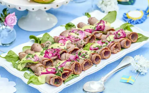 Rullad rostbiff med grön sås och rödlök, perfekt som enkel buffémat till många. Serveras på en vit bricka med gröna blad.