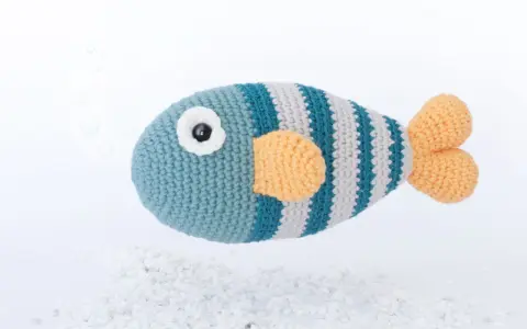 Un pez amigurumi a rayas azules y grises con aletas naranjas, flotando sobre guijarros blancos. ¡Encuentra aquí peces amigurumi patrón gratis!