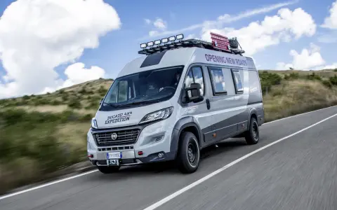 Szary kamper Ducato 4x4 Expedition z bagażnikiem dachowym i dodatkowym oświetleniem jedzie po drodze wśród wzgórz.