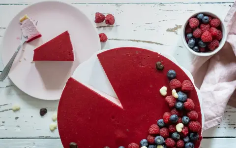 Ein köstlicher no bake Himbeer Cheesecake, dekoriert mit frischen Beeren und Schokoharz. Ein Stück ist bereits auf einem Teller.