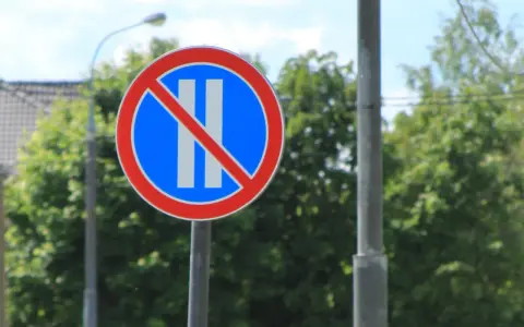 Znak B38 zakazujący postoju. Okrągły znak z czerwonym obramowaniem, niebieskim tłem i dwoma białymi pionowymi pasami przekreślonymi czerwoną linią.