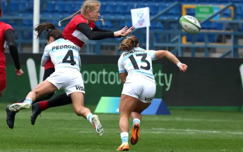 Zacięta walka o piłkę podczas rugby mistrzostwa świata. Zawodniczka w czerwonej koszulce próbuje przechwycić piłkę, podczas gdy dwie rywalki w białych strojach próbują ją zatrzymać.