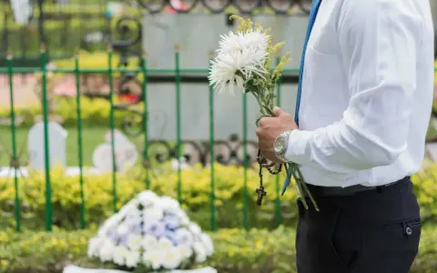 Mężczyzna w białej koszuli i niebieskim krawacie trzyma bukiet białych kwiatów i różaniec. To jego urlop okolicznościowy śmierć bliskiej osoby.