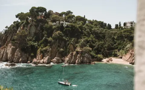 A sailboat floats in turquoise water near a rocky, tree-covered cliff with buildings. Perfect for weekend trips from Barcelona by train.