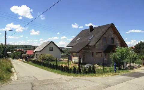 Dwa domy w budowie, jeden gotowy, na tle błękitnego nieba z chmurami. Widać drogę i zieleń. Różne kategorie gleby widoczne na poboczu.