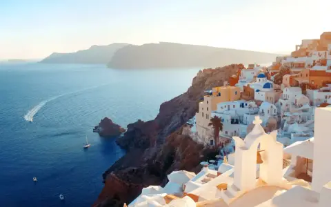Traumhafter Blick auf die weißen Häuser und blauen Kuppeln von Santorini. Das Meer glitzert, Boote fahren vorbei. Ein perfekter Santorini Strand Urlaub.