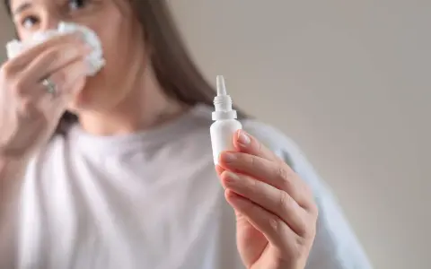 Kobieta z chusteczką przy nosie trzyma buteleczkę z kroplami do nosa, przygotowując się do plukania zatok.