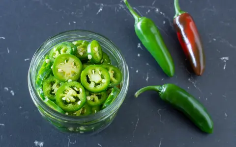 Frisch geschnittene Jalapeños in einem Glas, bereit zum Jalapenos einlegen. Daneben liegen drei ganze Chilis, eine davon reif rot.
