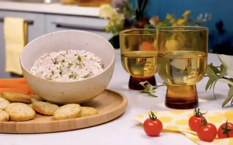 Un bol de rillettes de thon crémeuses, garni de ciboulette, servi avec des crackers, des bâtonnets de carottes et de céleri. Deux verres de vin blanc complètent cette scène gourmande.
