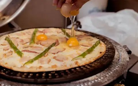 Ręka wylewa jajko na pizzę z boczkiem i szparagami. Pyszna pizza z jajkiem w przygotowaniu.
