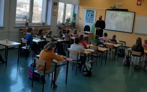 Dzieci słuchają lekcji o bezpieczeństwie w szkole. Nauczycielka i policjant prowadzą zajęcia.
