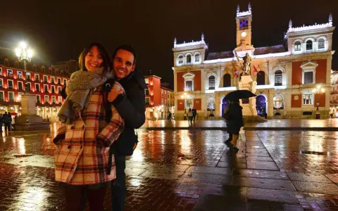Pareja sonriente en la Plaza Mayor de Valladolid, disfrutando de las atracciones nocturnas y el reflejo de las luces en el suelo mojado.