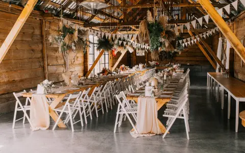 Drewniana stodoła udekorowana na przyjęcie. Długie stoły z kwiatami i świecami, wiszące dekoracje z traw. Widać różnicę między stylem boho chic a rustykalnym w dekoracjach kwiatowych.