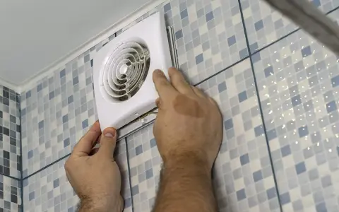 Mains d'un homme installant un ventilateur, une étape clé pour une bonne installation VMC dans une salle de bain carrelée.