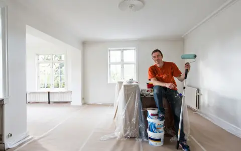Man med färgroller och hinkar redo att måla bakom elementet i ett rum under renovering.