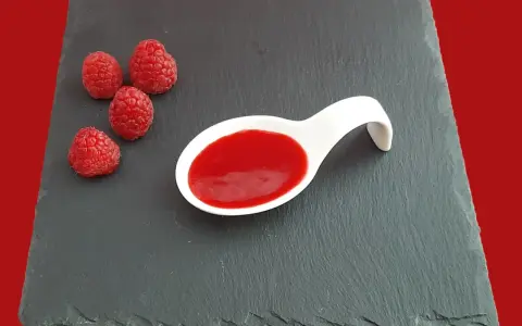Quatre framboises fraîches à côté d'une cuillère blanche remplie de coulis de framboise, sur une ardoise.