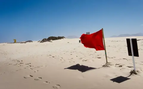 Pusta plaża z piaskiem, na którym widać ślady stóp. W oddali żółta budka ratownicza, a na pierwszym planie powiewają czerwone flagi ostrzegawcze.