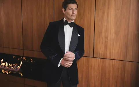 A man in a sophisticated navy velvet tuxedo, embodying perfect black tie attire, stands by a modern fireplace.