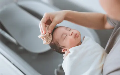 Bain d'un nouveau-né : une main nettoie délicatement la tête du bébé avec une petite serviette. Une scène paisible qui rappelle la liste produits toilette bébé naissance.