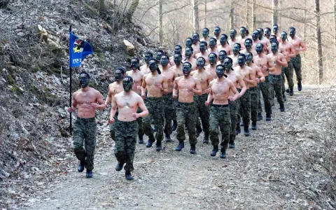 Żołnierze w kamuflażu biegają w lesie, niektórzy z zielonym malowaniem twarzy i flagą.