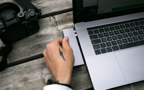 Dłoń z tatuażem pioruna podłącza adapter do laptopa, obok leży aparat. Idealny zestaw, jaki laptop do obróbki zdjęć?