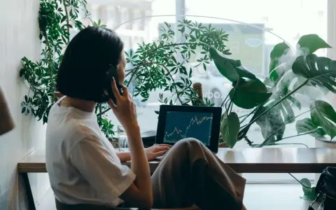 Kobieta w białej koszulce rozmawia przez telefon, pracując na laptopie z wykresem giełdowym. To jej wolny zawód, który pozwala jej pracować w otoczeniu zieleni.