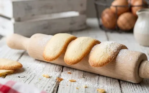 Biscotti a forma di lingue di gatto, spolverati di zucchero a velo, adagiati su un matterello di legno.