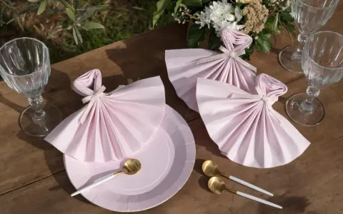 Vackert dukat bord med rosa servetter vikta som klänningar, guldfärgade skedar och glas. En bukett blommor fulländar dukningen.