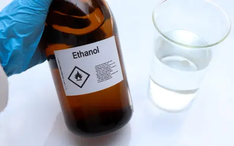 Braune Glasflasche mit der Aufschrift "Ethanol" und einem Warnsymbol. Daneben ein Becherglas mit klarer Flüssigkeit. Hier gibt es 100 prozentigen Alkohol.
