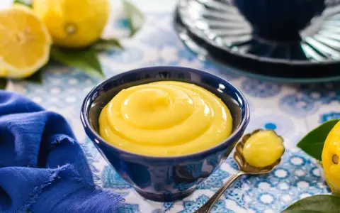 Una ciotola blu piena di vellutata crema al limone, con limoni freschi e un tovagliolo blu sullo sfondo.