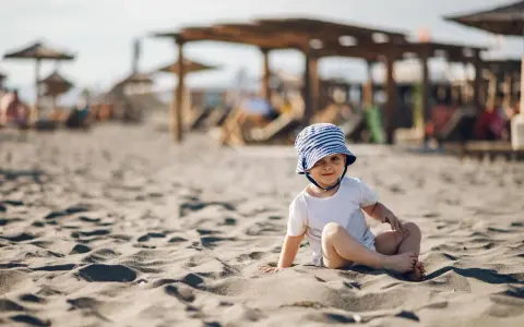 Uroczy maluch w pasiastym kapelusiku siedzi na piaszczystej plaży. Idealne miejsce, gdzie na wakacje z rocznym dzieckiem.