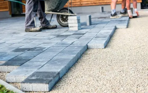 Układanie nowoczesnych płyt betonowych, co zamiast kostki brukowej. Pracownicy z taczką tworzą ścieżkę.