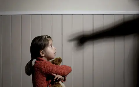 Mała dziewczynka z pluszowym misiem patrzy z przerażeniem na cień ręki wskazującej palcem, symbolizujący agresję wobec dziecka.