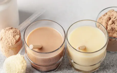 Dwa szklanki z napojami proteinowymi, obok proszek białkowy w miarkach i miseczkach.