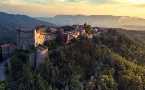 Antico castello medievale su una collina boscosa, uno dei tanti castelli da visitare in Toscana, con un villaggio arroccato e vista sulla valle al tramonto.