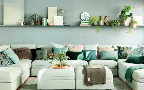 Sofá modular claro con cojines verdes y estampados, y una estantería con plantas y objetos decorativos, incluyendo una casita de cristal con **muebles pintados en verde**.