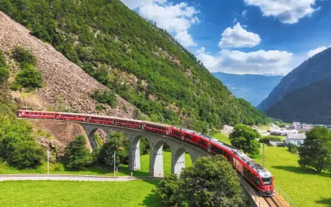 Czerwony pociąg Bernina Express przejeżdża przez kamienny wiadukt w Alpach. Sprawdź bernina express cena, by zaplanować podróż.