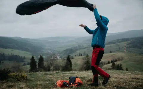 Osoba w niebieskiej kurtce i czerwonych spodniach rozkłada śpiwór puchowy czy syntetyczny na tle górskiego krajobrazu.