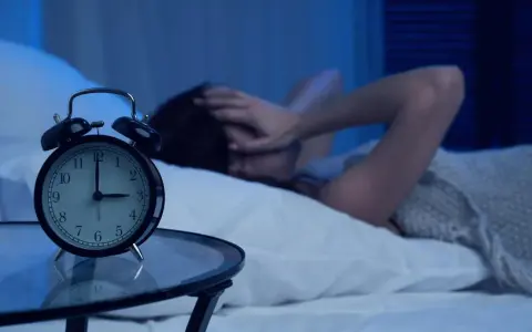 Eine Person liegt im Bett, die Hände über dem Gesicht, während ein Wecker auf 4 Uhr steht. Warum wache ich immer um 4 Uhr auf?