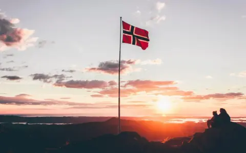 Zachód słońca w Norwegii. Dwie osoby siedzą na skale, patrząc na krajobraz. Flaga Norwegii powiewa na maszcie. Czy do Norwegii można lecieć na dowód osobisty?
