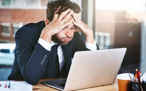Zestresowany mężczyzna w garniturze z rękami na głowie, patrzący na laptopa. Trudna spłata długu po ogłoszeniu upadłości.
