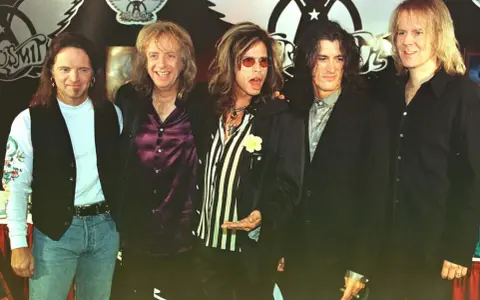 Steven Tyler mlody z zespołem Aerosmith, w charakterystycznych okularach i stroju, gotowy na scenę.
