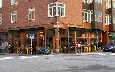 Ett mysigt hörnkafé i **Södermalm, Stockholm** med folk som sitter vid gula stolar och bord utomhus. En blå cykel med barnvagn står parkerad.
