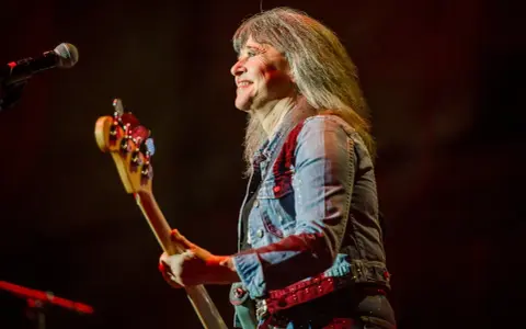 Suzi Quatro dzisiaj na scenie, z basem w ręku, uśmiecha się do publiczności. Rock'n'rollowa legenda w dżinsowej kurtce.