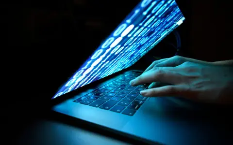 Händer på ett tangentbord, skärmen visar binär kod som flödar, en visuell representation av ett cyberattack idag.