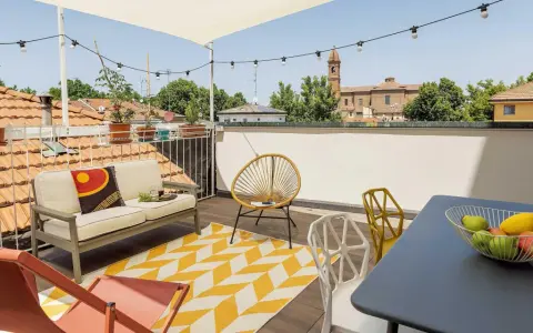 Terrazza sul tetto idee: divano, sedie colorate, tappeto giallo e bianco, tavolino con frutta, luci e vista sui tetti.
