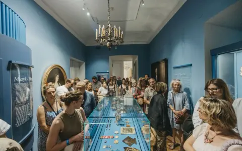Grupa zwiedzających ogląda wystawę w muzeum. Wśród eksponatów w gablocie widać ozdobne przedmioty, a na ścianach obrazy i tablice informacyjne. Klasyczny wystrój sali z żyrandolem stanowi tło dla tego współczesnego zainteresowania historią.