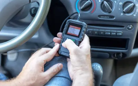 Ręce trzymające skaner diagnostyczny, który pokazuje menu z opcją "02 Sensor Test". To narzędzie pomoże zdiagnozować problemy z czujnikiem spalania stukowego.