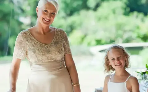 Elegancka stylizacja na komunię dla babci. Uśmiechnięta kobieta w beżowej sukni z haftem i dziewczynka w bieli.