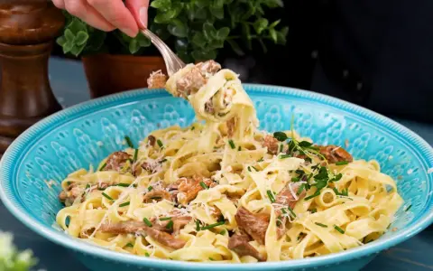 En gaffel lyfter en portion krämig pasta med kantareller, toppad med parmesan och färska örter i en vacker blå skål.